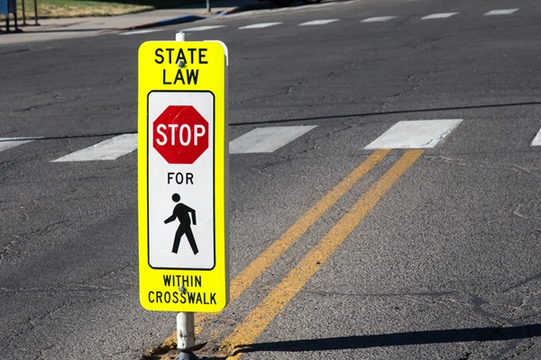Yellow crosswalk sign reading “State Law Stop for Pedestrians Within Crosswalk” positioned in the middle of a road.