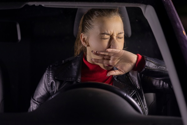 A female driver, seen at night, covering her mouth with her hand and yawning while driving, indicating extreme drowsiness.