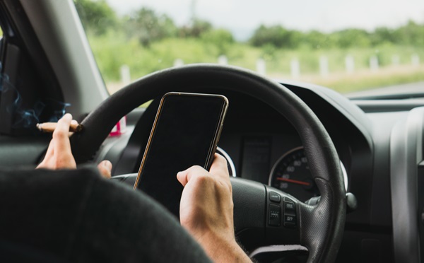 A person in the driver's seat of a car holding a smartphone and a lit, smoking marijuana cigar simultaneously.