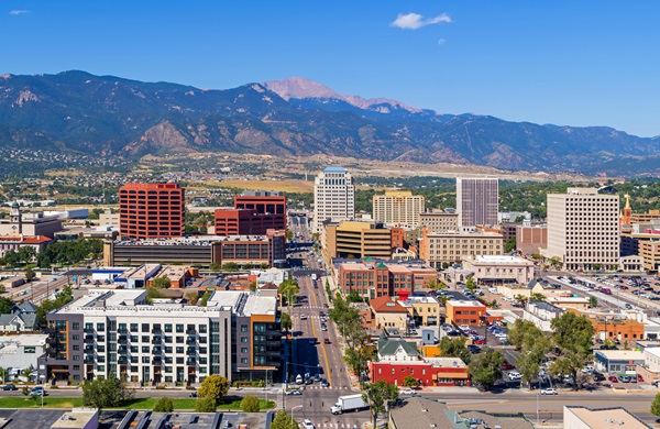 An aerial, high-angle view of downtown Colorado Springs, showing a mix of modern office buildings and low-rise urban structures under a bright blue sky, with the majestic, snow-dusted peaks of the Rocky Mountains rising in the background.