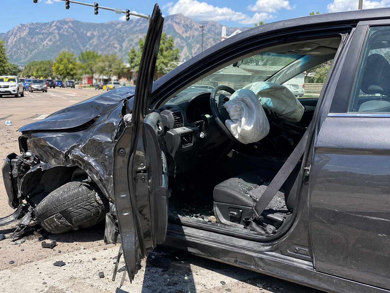 A dark sedan sits heavily damaged at a sunny intersection in Colorado Springs with its front end crushed, driver's side door open, and both front airbags deployed, revealing an unbuckled seat belt hanging over shattered glass on the interior seats against a backdrop of distant mountains.
