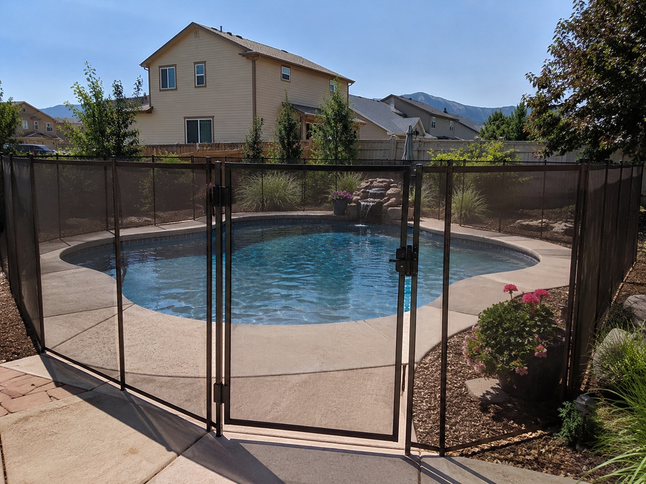 A round residential swimming pool in a Colorado suburban backyard is enclosed by a black mesh safety fence with a latched gate, with a rock waterfall feature, flowering plants, and the Rocky Mountains visible in the background, depicting the type of pool enclosure that Colorado property owners are required to maintain to reduce drowning risks and limit premises liability exposure under Colorado's attractive nuisance doctrine.